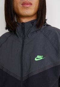 Črna in siva jakna na zadrgo s kapuco, ki ima na prsih zeleni znak Nike in teksturirano tkanino.