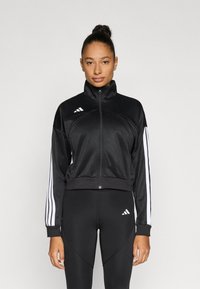 Črna športna jakna iz gladkega blaga, z visokim ovratnikom, zadrgo po celotni dolžini in belimi tremi črtami na rokavih.