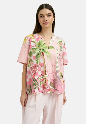 Woman wearing a loose pink short-sleeve blouse with tropical and beach-themed illustrations and light pink wide-leg pants.