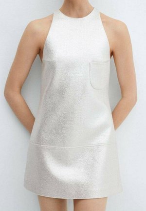 Frau trägt ein ärmelloses, hochgeschlossenes, elfenbeinfarbenes, strukturiertes Minikleid mit einer kleinen Brusttasche, steht vor einem schlichten hellen Hintergrund.