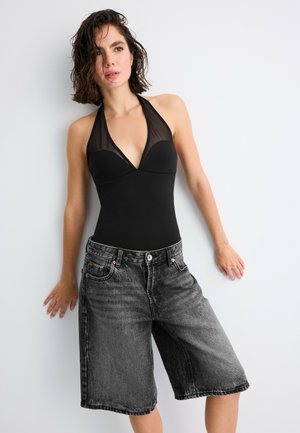 Femme aux cheveux courts et foncés, portant un top noir à col halter et un short en jean noir délavé, ample, debout contre un mur blanc.