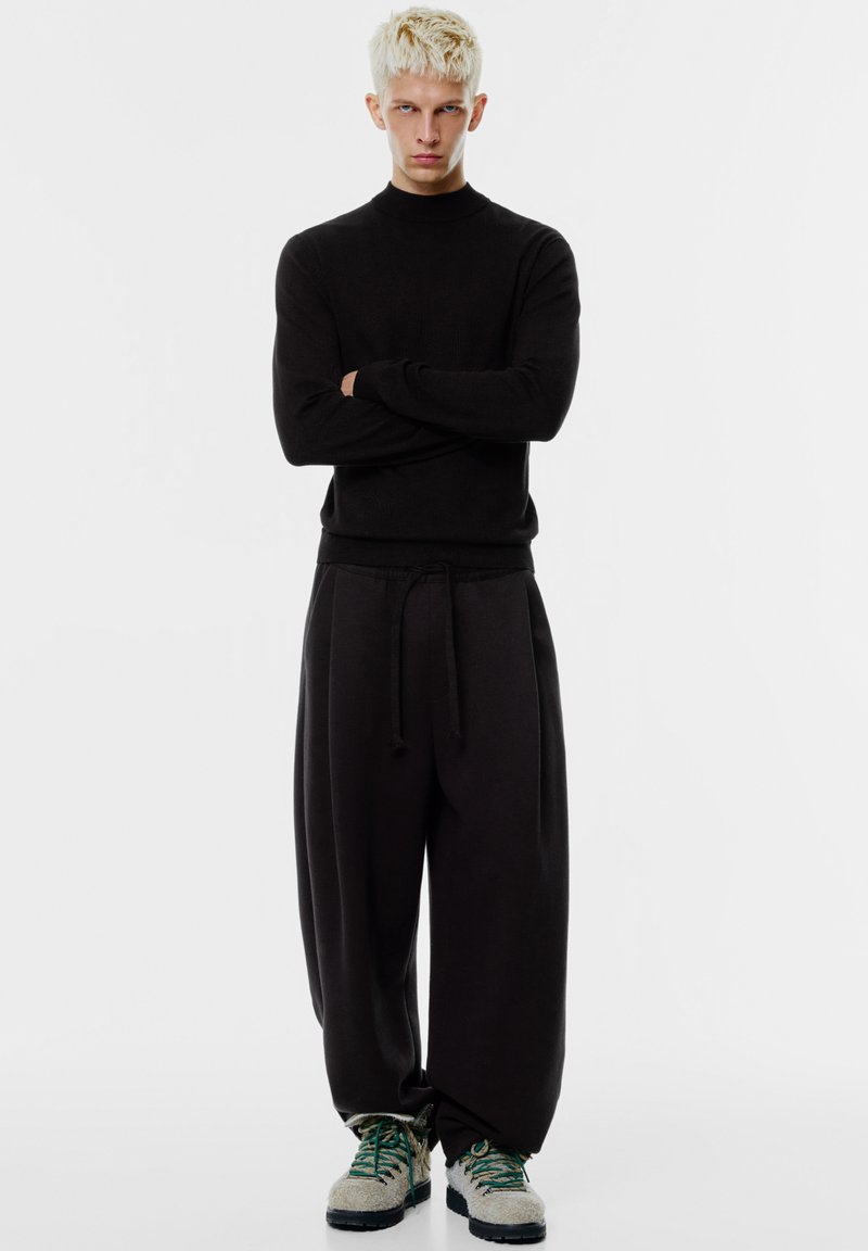 Svart figurnära tröja med hög hals, kombinerad med lösa svarta byxor med dragsko. Skorna är grå med gröna detaljer.