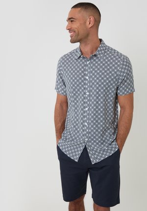 Mężczyzna uśmiechający się, ubrany w białą koszulę z krótkim rękawem z granatowym geometrycznym wzorem oraz ciemnogranatowe szorty, ręce w kieszeniach na jednolitym tle.