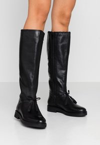 Bottes montantes en cuir noir avec une finition texturée, une fermeture éclair latérale et des pompons décoratifs sur les liens ajustables à la cheville.