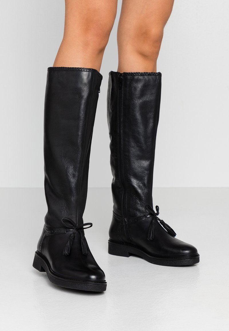 Bottes montantes en cuir noir avec une finition texturée, une fermeture éclair latérale et des pompons décoratifs sur les liens ajustables à la cheville.