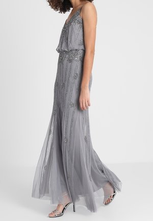 Vrouw die loopt in een grijze jurk tot op de grond, zonder mouwen, met kralenversiering en zilveren sandalen met hoge hakken.