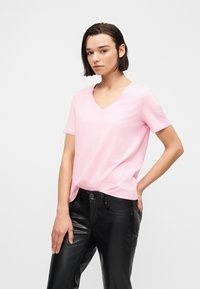 Νέα γυναίκα με κοντά σκούρα μαλλιά, φορώντας ανοιχτό ροζ T-shirt με V λαιμόκοψη και μαύρο δερμάτινο παντελόνι, στέκεται μπροστά σε απλό φόντο.