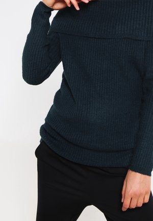 Person iført en mørkeblå ribstrikket sweater og sorte bukser, med den ene hånd, der rører ved sweaterens krave.