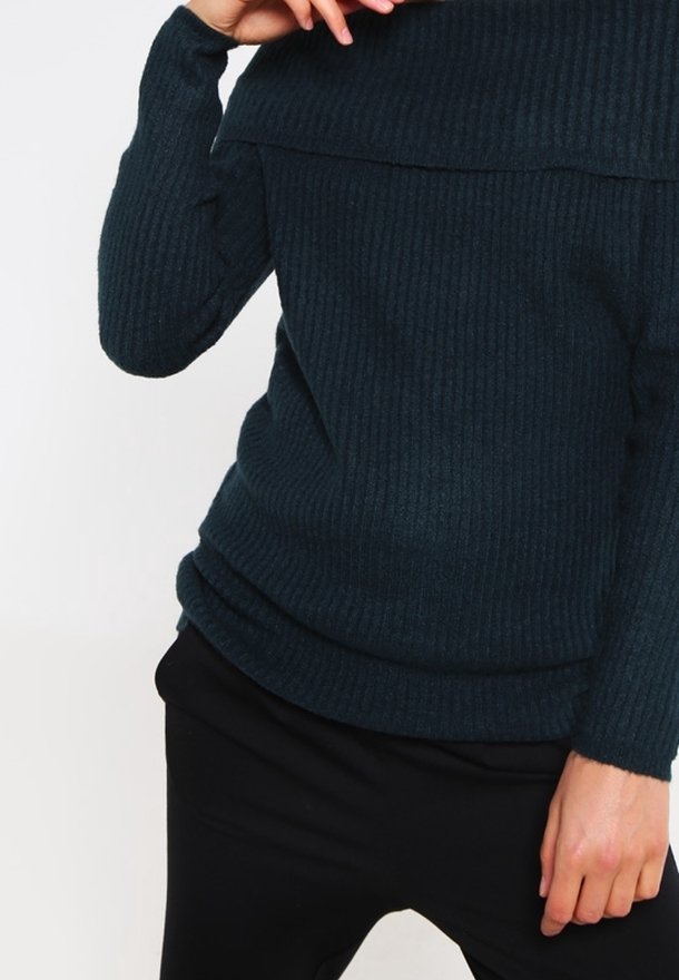 Personne portant un pull côtelé bleu foncé et un pantalon noir, une main touchant le col du pull.
