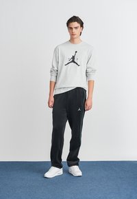 Hanorac gri deschis cu logo Jumpman negru, asortat cu pantaloni de trening negri, având o fâșie albastră și logo, completat cu adidași albi.