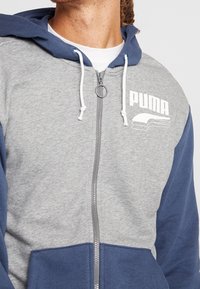 Hoodie zippé gris et bleu marine avec un logo PUMA blanc, doté d'une capuche à cordon de serrage et d'une finition texturée. Les détails comprennent une fermeture éclair.