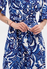 Sukienka z krótkim rękawem w kolorze białym z niebieskim kwiatowym wzorem, z paskiem w talii i lekko opadającym krojem. Gładka faktura i żywy kontrast kolorów.