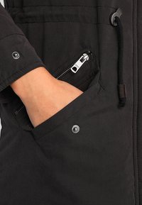 Main dans une poche de veste noire avec fermeture éclair et détails à bouton pression, et un cordon noir visible sur le côté.