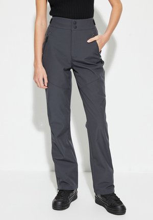 Femme portant un pantalon taille haute gris foncé avec des poches zippées et des chaussures noires, se tenant avec une main dans la poche sur un fond blanc.