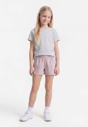 Ung pige med langt blondt hår iført en grå t-shirt, lyserøde shorts, hvide sokker og hvide sneakers, stående med et let smil.