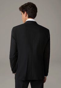 Giacca nera vista da dietro, con una texture liscia, spalle strutturate e uno spacco singolo, completata da un colletto di camicia bianco.