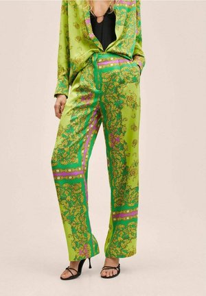 Vrouw draagt heldergroene wijde broekspijpen en een bijpassend colbert met gouden en paarse barokke patronen, gecombineerd met zwarte sandalen met bandjes.