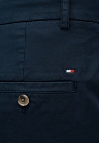 Παντελόνι ναυτικό μπλε με λεία υφή, διαθέτει πλαϊνή τσέπη και ορατό καφέ κουμπί· περιλαμβάνει ένα μικρό λογότυπο σε κόκκινο, λευκό και μπλε.