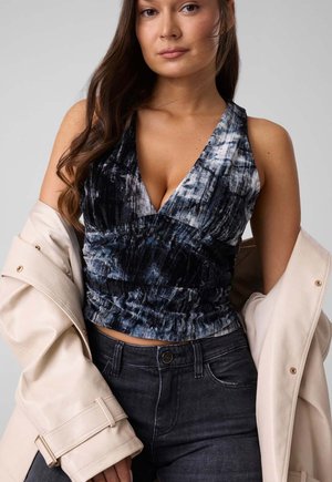 Joven mujer con una blusa azul sin mangas con estampado, vaqueros oscuros, y sosteniendo una chaqueta beige sobre sus brazos.