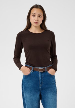 Jeune femme aux longs cheveux bruns porte un haut moulant à manches longues marron foncé, un jean bleu et une ceinture marron, debout les mains dans les poches.