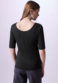 Svart figurnära topp med bred rundad halsringning och korta ärmar, tillverkad av jersey-material. Baksidan har en låg skärning och har en slät yta.