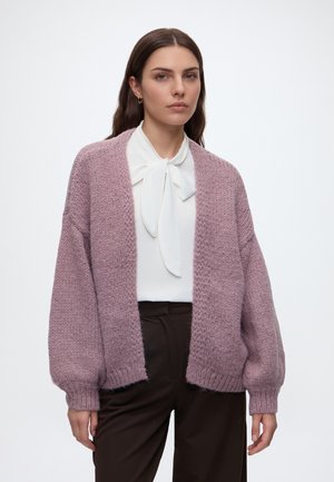 Femme portant un chemisier blanc avec un nœud noué et un cardigan mauve texturé et ample sur un pantalon marron foncé devant un fond clair uni.