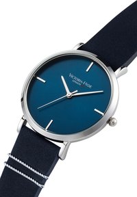 Montre en acier inoxydable argenté avec un cadran bleu foncé, dotée d'index et d'aiguilles métalliques, et d'un bracelet en tissu bleu marine avec des accents argentés.