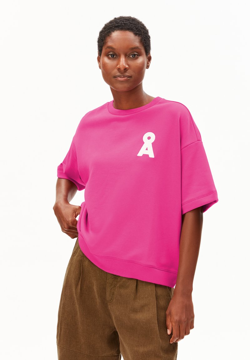 Frau trägt ein leuchtend pinkes, übergroßes Hemd mit weißem Å-Logo auf der Brust und lockere braune Hose, steht vor weißem Hintergrund.
