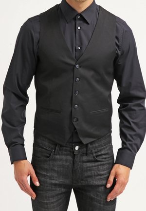 Mężczyzna ubrany w dopasowaną czarną kamizelkę na guziki, na czarnej koszuli z długim rękawem, w ciemnoszarych jeansach, stojący z rękami rozluźnionymi.