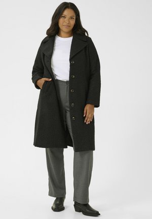 Black textured coat with a notched collar, button-front closure, and side pockets. Worn over gray trousers and black ankle boots.