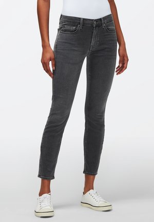 Person iført slim-fit mørkegrå jeans og hvide sneakers, stående mod en ensartet lys baggrund.