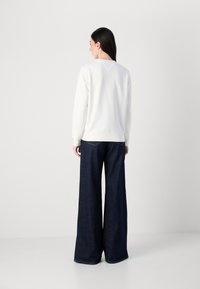 En kvinne står med ryggen til betrakteren, iført en hvit genser og vide, mørke jeans mot en lys grå bakgrunn.