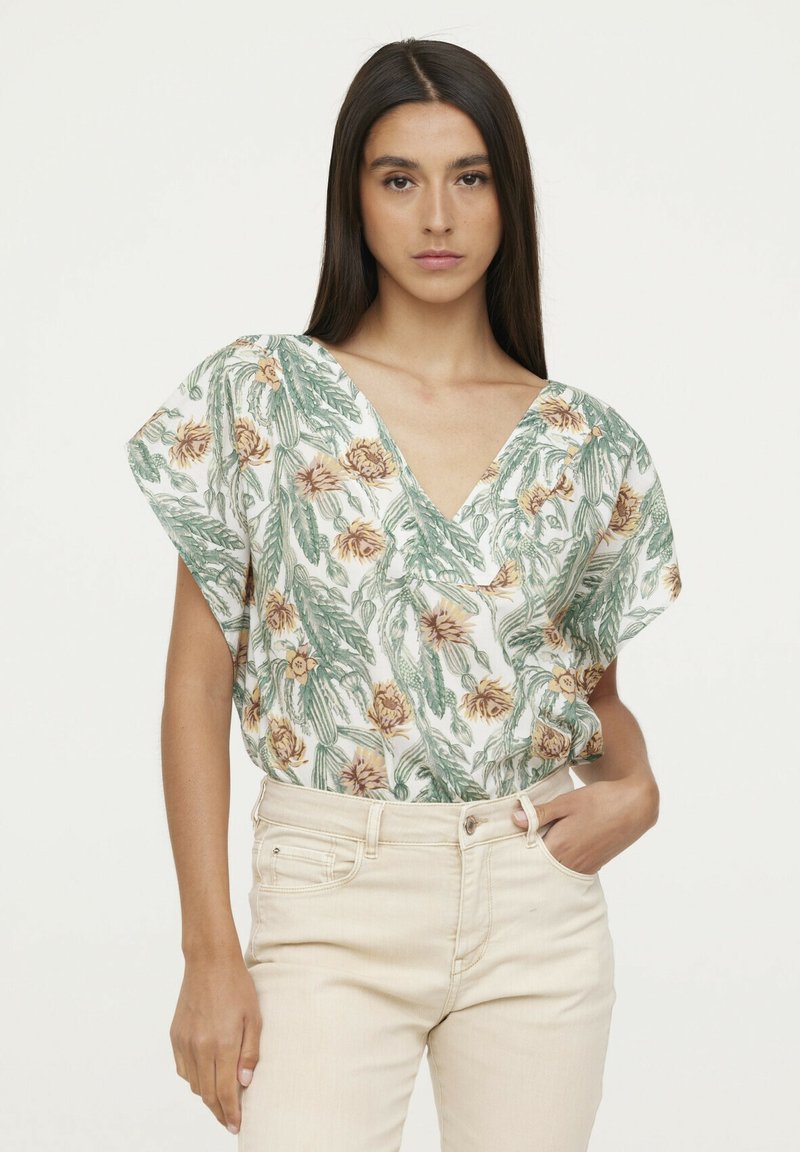 Blouse florale avec un décolleté en V, des manches courtes, et un motif de feuilles vertes et de fleurs jaunes sur fond blanc. Associée à un jean clair.