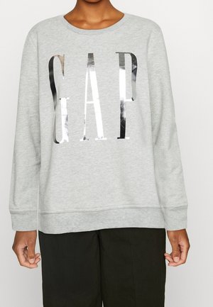 Osoba ubrana w jasnoszarą bluzę z dużymi metalicznymi srebrnymi literami "GAP" na przodzie oraz czarne spodnie, stojąca na tle jednolitego koloru.