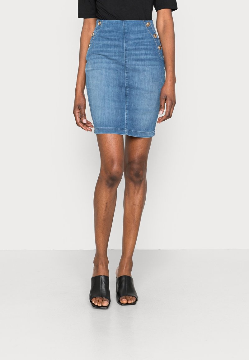 Minifalda de mezclilla en azul claro con un diseño ajustado, cintura alta y detalles de botones en los lados. Unos zapatos de slide negros completan el look.