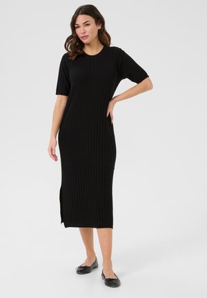 Black ribbed knit dress with short sleeves, knee-length, featuring side slit. Paired with black flat shoes, set against a neutral background.