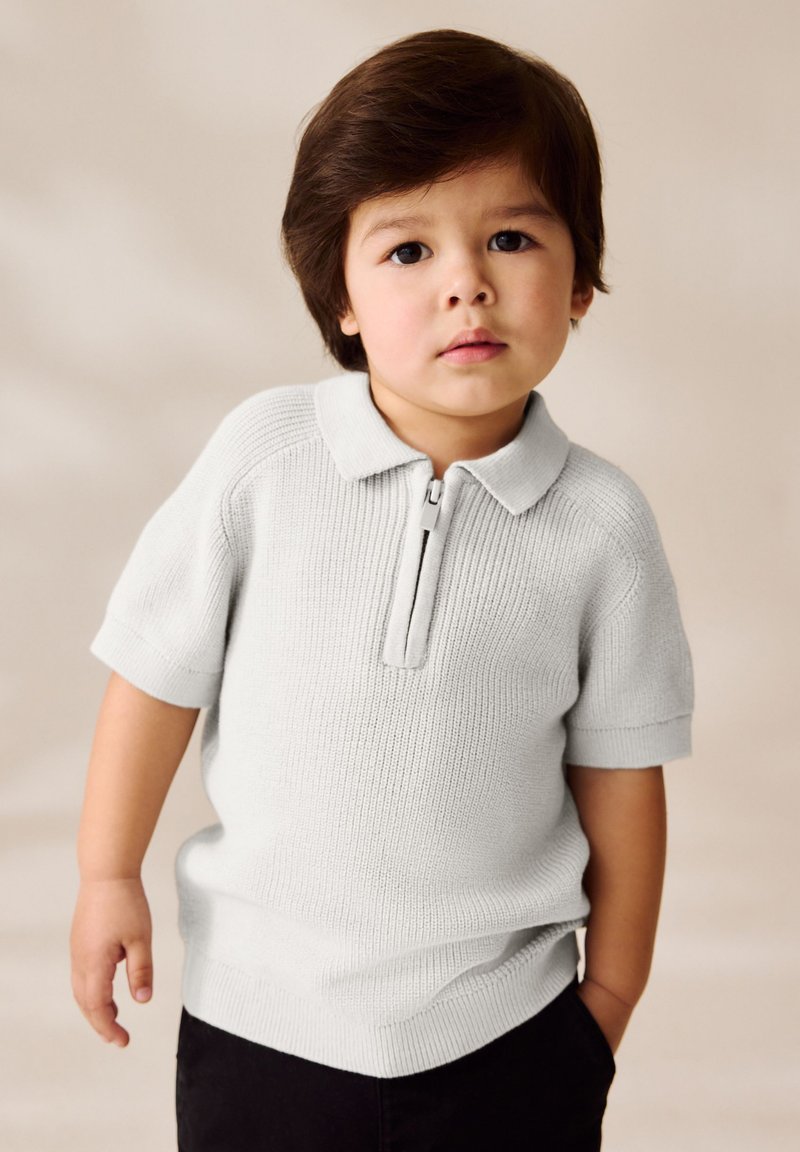 Kleines Kind mit dunklen Haaren trägt ein hellgraues, kurzärmliges Strick-Poloshirt und schwarze Hosen und steht vor einem beigefarbenen Hintergrund.