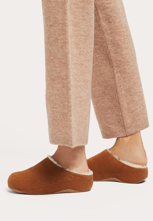 Braune Wildlederschlappen mit einem flauschigen weißen Futter und einem niedrigen Rückendesign, kombiniert mit hellbeigen, leicht strukturierten Loungehosen.