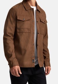 Veste marron texturée à boutons, avec deux poches avant, boutons-pression et col pointu. Portée par-dessus une chemise blanche à manches longues.
