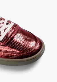 Zapatilla deportiva metálica roja con acabado texturizado, cordones blancos y suela de goma gris contrastante. Punta redondeada con lengüeta acolchada.