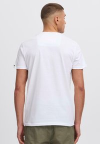 Camiseta blanca de algodón con mangas cortas, cuello redondo y un parche rectangular en la parte superior de la espalda. Textura suave, corte relajado.
