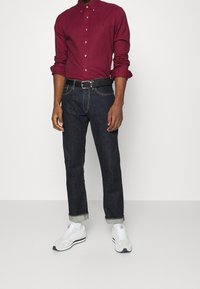 Ruitjesoverhemd in rood en wit, donkerblauwe denimjeans, zwarte riem, opgerolde mouwen en witte sneakers met grijze accenten.