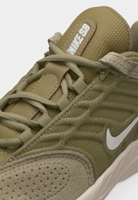 Κοντινό πλάνο αθλητικού παπουτσιού Nike SB σε ελιά πράσινο, που δείχνει υφασμάτινη υφή, κορδόνια, λευκό λογότυπο swoosh και ετικέτα με λογότυπο στη γλώσσα.