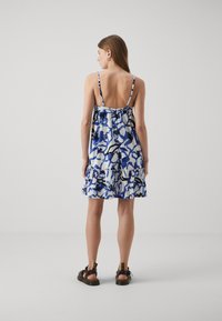 Rochie de soare cu model albastru și alb, cu bretele subțiri și un tiv ondulat. Are un spate decupat și o croială lejeră. Este purtată cu sandale maro închis.
