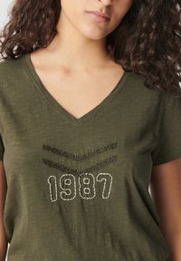 Camiseta verde oliva con cuello en V, que presenta una costura texturada en negro y blanco en patrón de espiga y el número "1987" destacado.