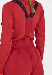 Röd långärmad topp med texturerad tyg, med hög krage och svarta justerbara remmar. Matchande högmidjade byxor med elastisk midja.