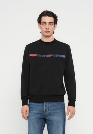 Čierna mikina s okrúhlym výstrihom a rebrovanými manžetami; na prednej strane potlačené logo v červenej a modrej farbe. Oblečená s modrými rifľami.