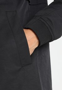 Main insérée dans la poche latérale d'une veste noire avec un poignet côtelé sur la manche, vue en gros plan.