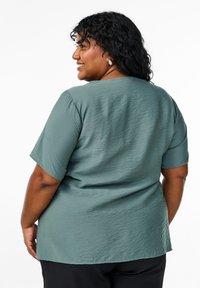 Top teal a maniche corte con un design testurizzato e increspato. Orlo leggermente curvo, scollatura rotonda e vestibilità rilassata. Tessuto semplice, senza motivi.