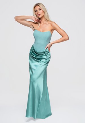 Mujer rubia posando con vestido de satén verde azulado sin tirantes, con corpiño ajustado y falda drapeada sobre fondo liso y claro.